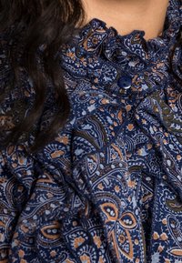 Blusa blu navy con scollatura arricciata, caratterizzata da un intricato motivo paisley in arancione e verde. Include un dettaglio di bottone blu.