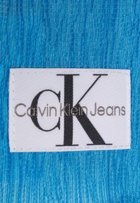 Etykieta Calvin Klein Jeans na teksturowanej niebieskiej tkaninie. Etykieta jest biała z czarnym napisem zawierającym "CK" oraz "Calvin Klein Jeans."