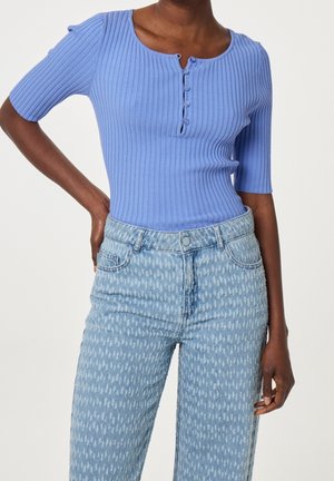 Kobieta w obcisłej, prążkowanej jasnoniebieskiej bluzce z guzikami z przodu oraz jasnoniebieskich, wzorzystych, wysokich dżinsach, stojąca z jedną ręką na biodrze.