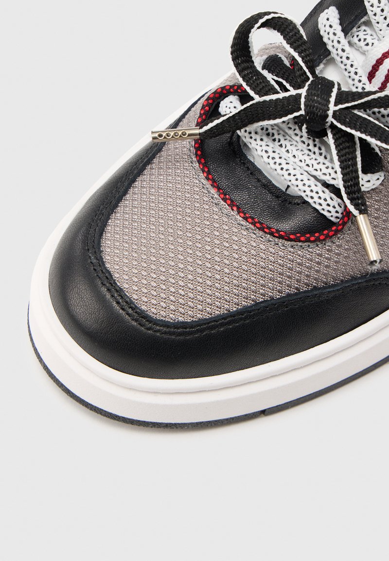 Sneakers en cuir noir et mesh gris avec semelle blanche, présentant des coutures rouges, une surface texturée et des lacets noirs avec des accents en métal sur les embouts.