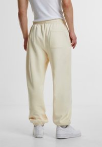 Persona vestida con pantalones de chándal color crema con tobillos y cintura elásticos, zapatillas blancas y una camiseta sin mangas blanca, de pie frente a un fondo blanco.