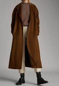 Cappotto di lana marrone con un ampio colletto, abbinato a un maglione di lana marrone e pantaloni cropped avorio; presenta stivali neri con lacci.
