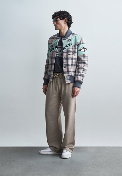 Homme aux cheveux bouclés portant des lunettes, une veste bomber à carreaux avec des accents menthe, un t-shirt graphique noir, un pantalon beige et des baskets blanches, debout de profil.