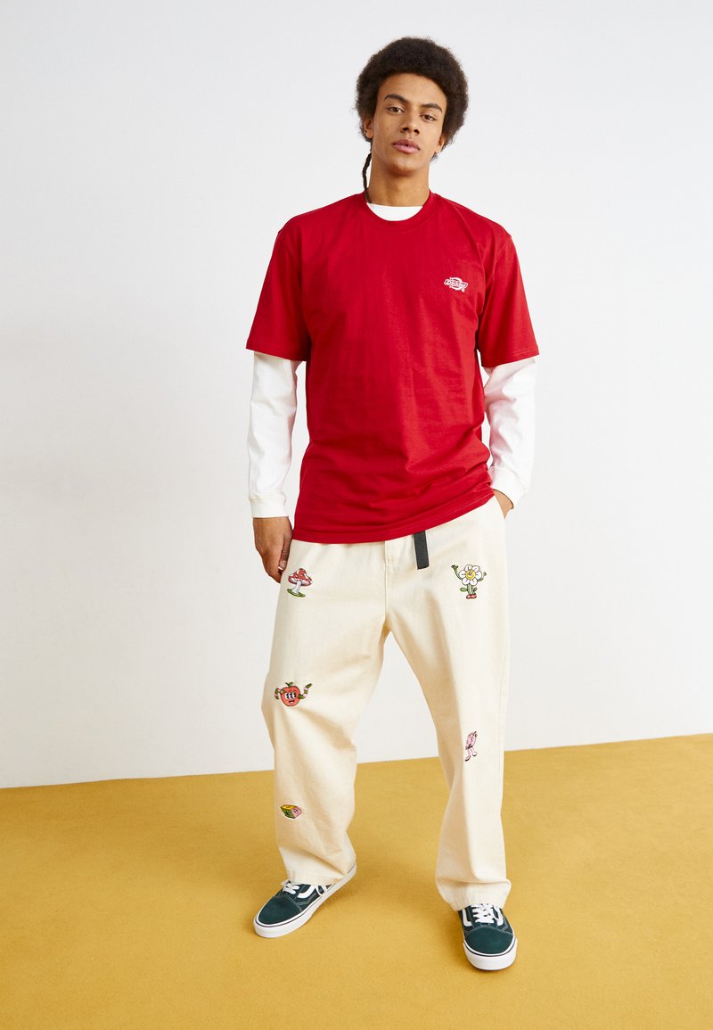 Rode katoenen T-shirt gelaagd over een witte lange mouwen shirt, gecombineerd met crèmekleurige broek met kleurrijke geborduurde fruit- en bloemontwerpen.