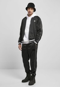 Giacca bomber nera con accenti bianchi, sovrapposta a una felpa bianca con cappuccio, abbinata a joggers neri con logo e stivali neri.