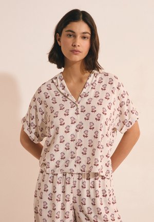 Młoda kobieta o krótkich ciemnych włosach, ubrana w biały kwiecisty top na guziki i luźne spodnie, stoi z rękami za plecami.