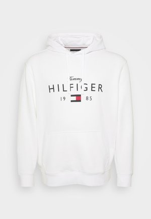Sweat à capuche blanc en tissu doux, doté d'une poche kangourou à l'avant. Un logo en texte noir et rouge indique "Tommy HILFIGER 1985" sur la poitrine.