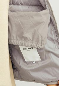 Jasnoszara ocieplana kurtka wewnątrz z miękką podszewką, z metką zawierającą napis "JACK & JONES" oraz informacje o rozmiarze.