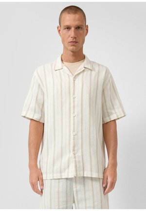 Mężczyzna ubrany w kremową koszulę z krótkim rękawem na guziki i dopasowane szorty z cienkimi pionowymi beżowymi paskami, stojący na jednolitym tle.