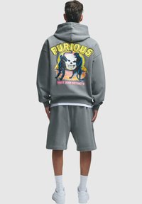 Grå oversized hettegenser med et levende grafisk motiv på ryggen, som viser et hodeskalle, bjørner og tekst. Sammen med matchende shorts og joggesko.