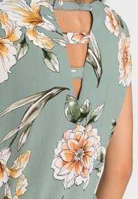 Haut fleuri en tissu vert clair avec des motifs de fleurs orange et blanches, un dos en V, et une petite sangle en tissu pour le détail.