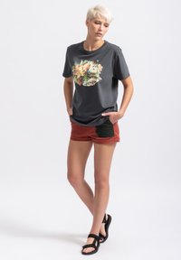 Grijze T-shirt met een kleurrijke bloemenafbeelding, gecombineerd met roestkleurige shorts met zwarte accenten. Model draagt zwarte sandalen.