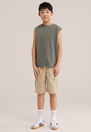 Jongen staat, draagt een mouwloos olijfgroen shirt, beige cargoshorts, witte sokken en witte sneakers met zwarte strepen.