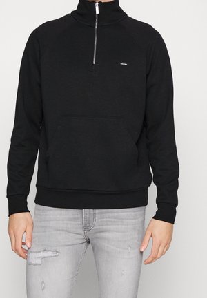 Mann trägt schwarzen Viertel-Zip-Sweatshirt mit Fronttasche und kleinem Logo, kombiniert mit hellgrauen, abgenutzten Jeans.