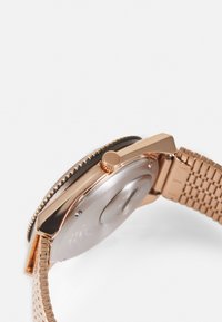 Roségoldene Uhr mit geflochtenem Metallarmband, rechteckigem Ziffernblatt und strukturiertem Gehäuse. Verfügt über eine Krone und ein poliertes Rückengehäuse.