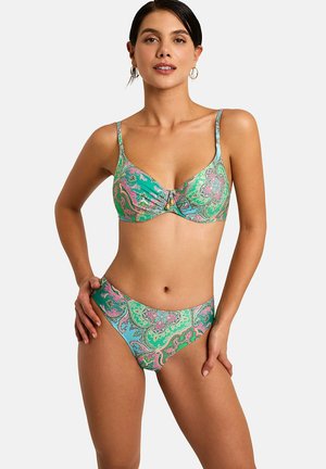 Vrouw draagt een bikini met een groene en blauwe paisleyprint en dunne bandjes, staat met één hand op de heup tegen een witte achtergrond.