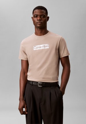 Uomo che indossa una t-shirt beige Calvin Klein infilata in pantaloni neri plissettati con cintura nera, in piedi davanti a uno sfondo grigio uniforme.
