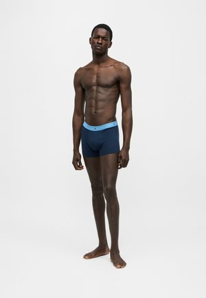 Modelo masculino en forma de pie descalzo vistiendo bóxers azul marino con cinturilla azul claro sobre fondo blanco liso.