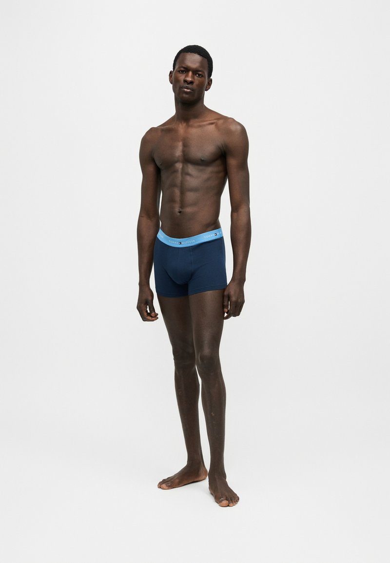 Λεπτός άνδρας μοντέλο στέκεται ξυπόλητος φορώντας μπλε μποξεράκια με ανοιχτό μπλε μέση σε απλό λευκό φόντο.