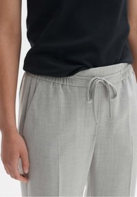 Pantalon gris clair, ample, avec une taille élastique et un cordon de serrage. Le tissu semble lisse avec une texture subtile.