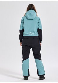 Een two-tone skibroek met een teal bovenstuk en een zwart onderstuk, met de merknaam "SLADE" op de achterkant.