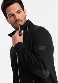Giacca nera con zip e collo alto, in tessuto testurizzato, con toppe alle elbow in similpelle liscia. Dotata di zip argentata e patch con logo.