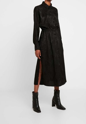 Black, long-sleeve shirt dress with textured pattern, button-up front, cinched waist, side slits, paired with black, heeled ankle boots.