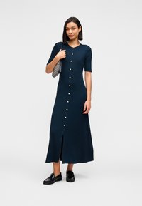 Πλεκτό φόρεμα σε σκούρο ναυτικό μπλε χρώμα με κοντά μανίκια, κουμπιά στο μπροστινό μέρος, ριμπ υφή και σκίσιμο μπροστά. Συνδυασμένο με μαύρα loafers και τσάντα με σχέδιο.