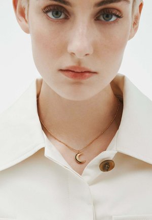 Collarín de camisa beige claro con detalles de botón, con un collar de luna creciente dorada con un acabado texturizado sobre una delicada cadena.