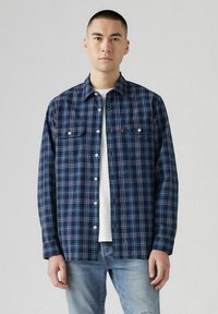 Camicia a quadri blu navy con accenti bianchi e blu, caratterizzata da due tasche sul petto, chiusura frontale con bottoni e vestibilità comoda. Indossata sopra una maglietta bianca.