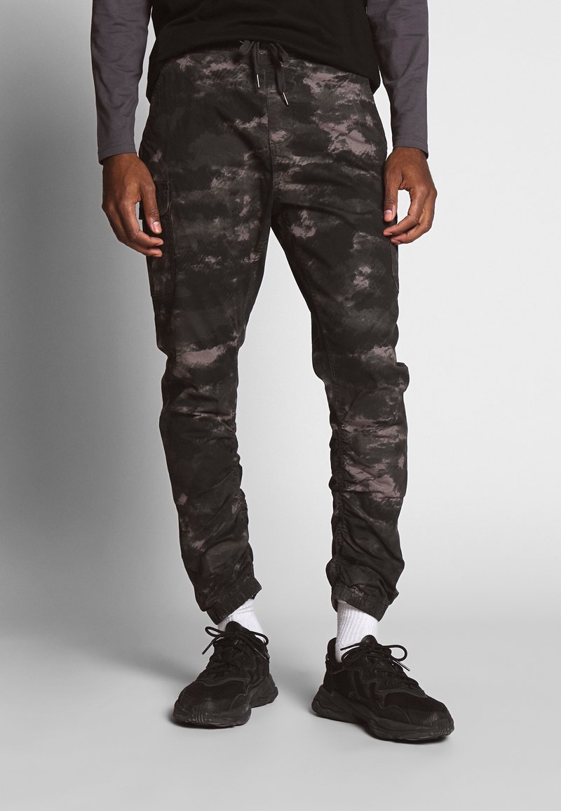 Personne portant un pantalon de jogging camouflage noir et gris, des baskets noires, des chaussettes blanches et une chemise à manches longues, debout devant un fond gris.