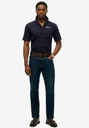 Camisa de manga corta navy con logo bordado, cuello abotonado, jeans oscuros, cinturón marrón y zapatos oscuros, de pie frente a un fondo sencillo.
