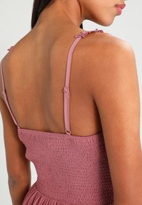 Vaaleanpunainen smokkimekko ohut säädettävillä olkaimilla. Koristeena herkät kukkakuvastot olkainten päässä ja virtaava hame. Sileä pinta.