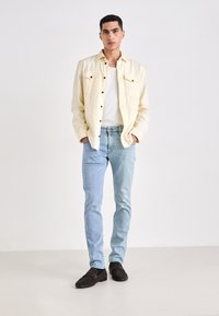 Világos sárga gombos ing két mellzsebbel, fehér ujjatlan póló, világoskék farmernadrág és fekete slip-on cipő, egy világos háttér előtt állva.