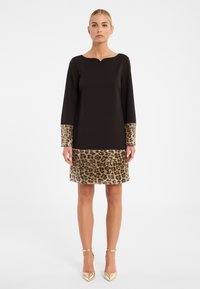 Schwarzes Kleid mit langen Ärmeln, gewelltem Halsetikett, Saum und Manschetten aus Leopardenmuster-Pailletten und einer glatten Textur. Hohe Absätze und flacher Hintergrund.