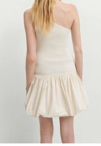 Femme aux longs cheveux blonds portant une robe crème sans manches en maille côtelée, avec une jupe bouffante froncée, vue de dos.