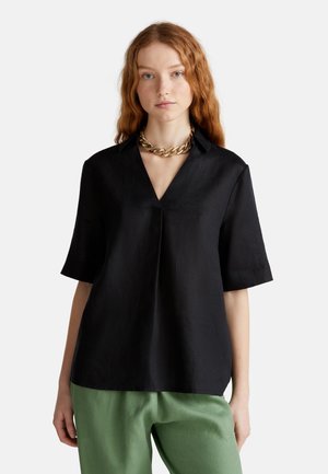 Zwarte top met korte mouwen en een V-hals, met een gladde textuur en een losse pasvorm, gecombineerd met groene broek. Accenten van een chunky gouden ketting.