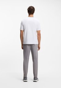 Άνδρας που στέκεται με την πλάτη γυρισμένη, φορώντας λευκό πουκάμισο με κοντά μανίκια, γκρι παντελόνι τύπου jogger και γκρι αθλητικά παπούτσια σε λευκό φόντο.