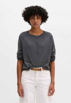 Femme aux cheveux bouclés portant un cardigan gris foncé boutonné, un pantalon blanc et une ceinture marron, debout devant un fond clair uni.