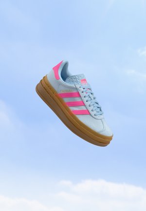 Basket Adidas Gazelle bleu clair avec des bandes roses et un détail au talon, flottant sur un fond de ciel clair.
