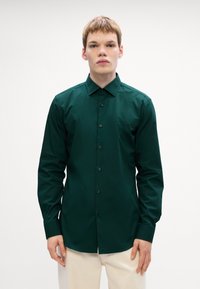 Donkergroene blouse met lange mouwen, een puntige kraag en zwarte knopen. Gladde, matte stof met een klassieke pasvorm.