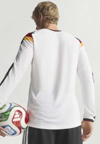 White long-sleeve sports jersey with black, red, and yellow accents on the shoulders; textured fabric; held football in the left hand.