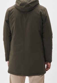 Man i olivgrön vinterjacka med huvtröja sedd bakifrån, beige byxor syns nedanför jackans nederkant.