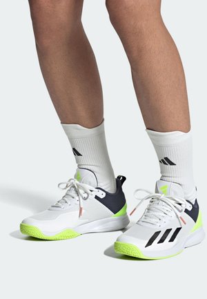 Des chaussures de sport blanches présentent un dessus en maille, des accents noirs et une semelle extérieure vibrant en vert. Portées avec des chaussettes blanches, montrant la texture et l'ajustement.