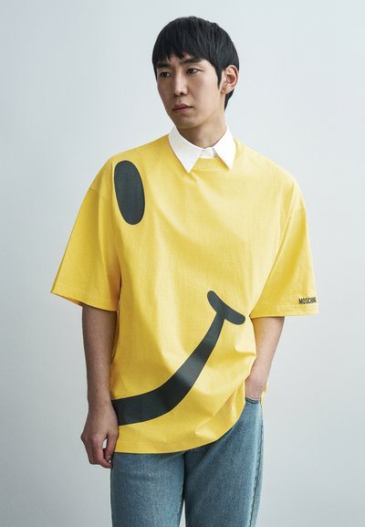 T-shirt jaune surdimensionné avec un grand graphique de smiley noir. Présente un col blanc et une étiquette portant l'inscription "MOSCHINO". Texture en coton lisse.