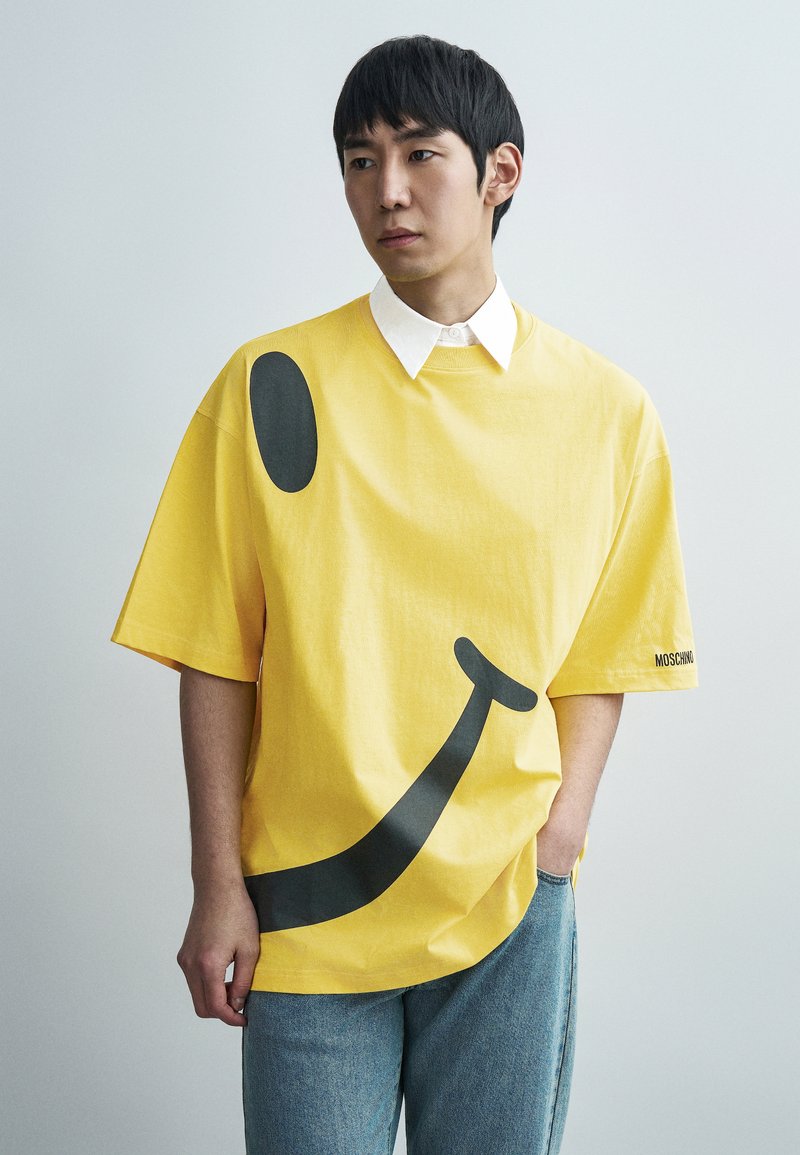 Κίτρινη μεγάλη κοντομάνικη μπλούζα με μεγάλο μαύρο γραφικό χαμογελαστό πρόσωπο. Διαθέτει λευκή γιακά και ετικέτα με την ένδειξη "MOSCHINO." Ομαλή υφή βαμβακιού.