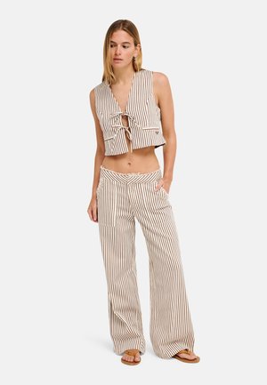 Femme portant un crop-top sans manches à rayures verticales beige et marron et un pantalon à jambes larges avec des sandales, debout sur un fond uni.