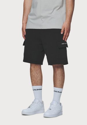 Man draagt zwarte cargoshorts met zijzakken, witte sportsokken en witte sneakers, staand tegen een effen achtergrond.