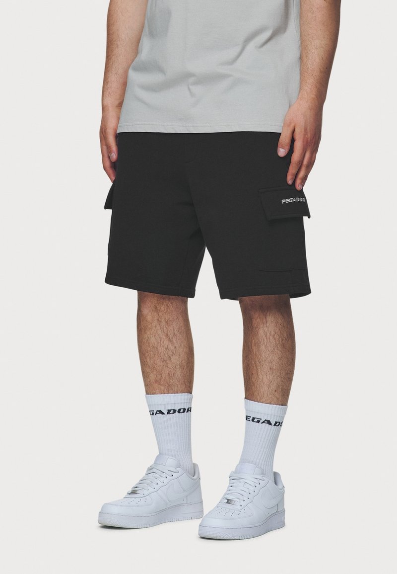 Homme portant un short cargo noir avec des poches latérales, des chaussettes blanches montantes et des baskets blanches, se tenant devant un fond uni.