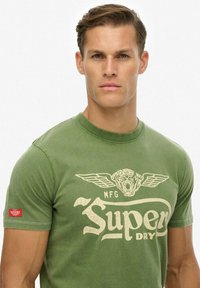 Camiseta de algodón verde con un gráfico beige de un tigre con alas y el texto "Super Dry" en un estilo vintage. Mangas cortas, cuello redondo.